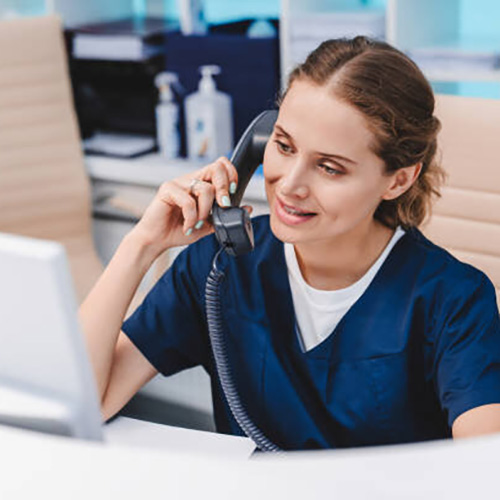 Young female receptionist talking on phone in clinic while sitting and looking on pc monitor